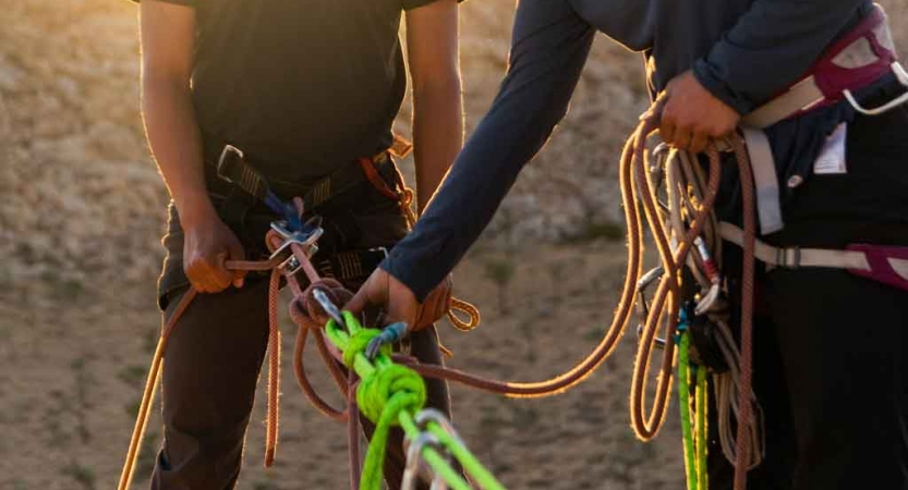 Two people wearing climbing gear stand near each other. One of them checks the ropes and knots of the other. 